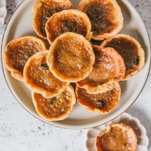 Butter tarts sit on a rimmed ceramic plate with one served up on an individual plate on a light gray surface.