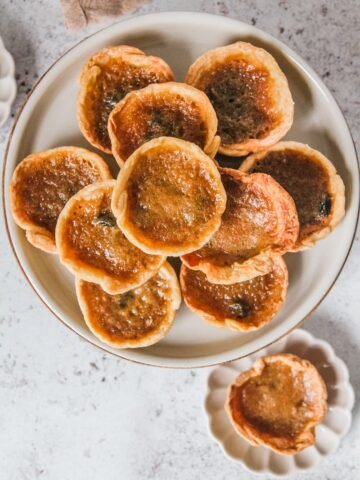 Butter tarts sit on a rimmed ceramic plate with one served up on an individual plate on a light gray surface.