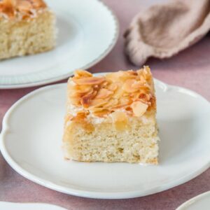 A slice of Butterkuchen sits on an individual white ceramic serving plate on a light pink surface.