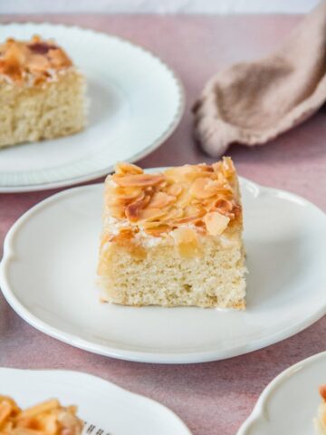 A slice of Butterkuchen sits on an individual white ceramic serving plate on a light pink surface.