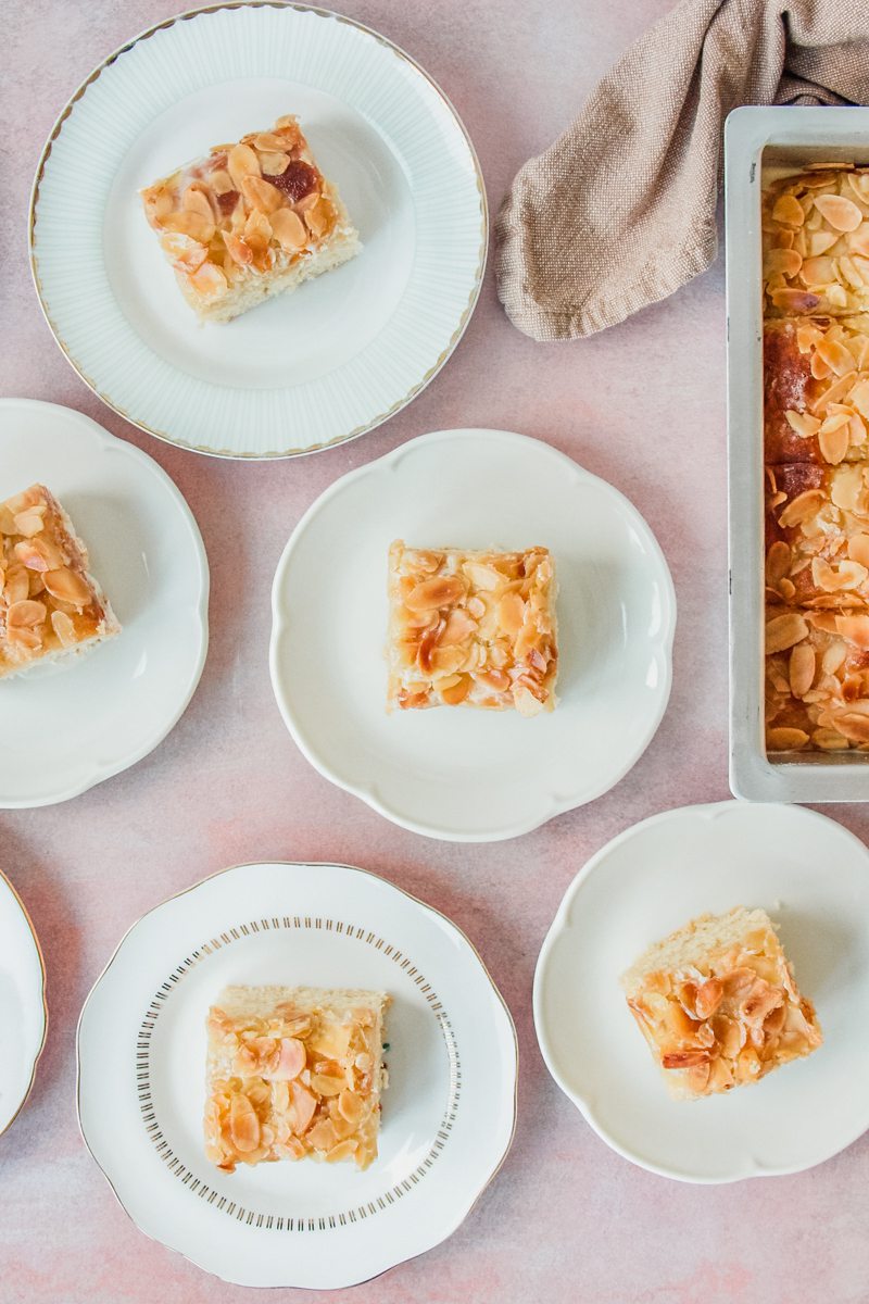 Slices of German Butter Cake sit on individual white ceramic plates on a light pink surface.