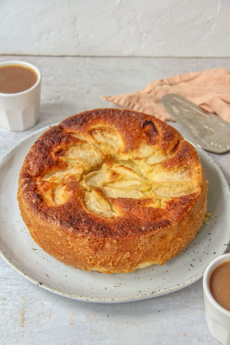 British buttery Dorset Apple Cake sits on a light blue ceramic plate on a grey surface with two small cups of coffee beside.