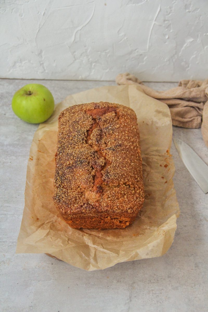 Golden apple butter cake sits on parchment paper on a gray surface with an apple and brown cloth behind.