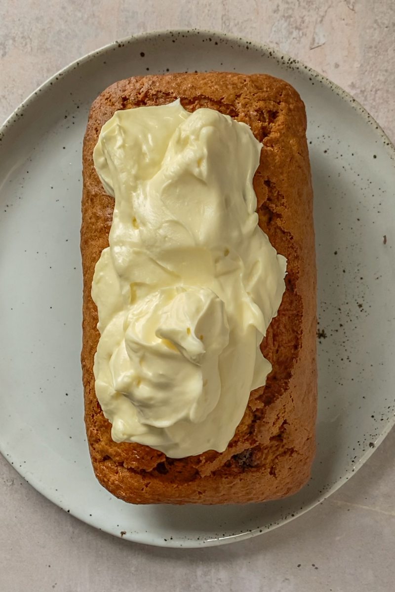 Cream cheese frosting sits on top of carrot cake loaf on a blue ceramic speckled plate on a gray surface.