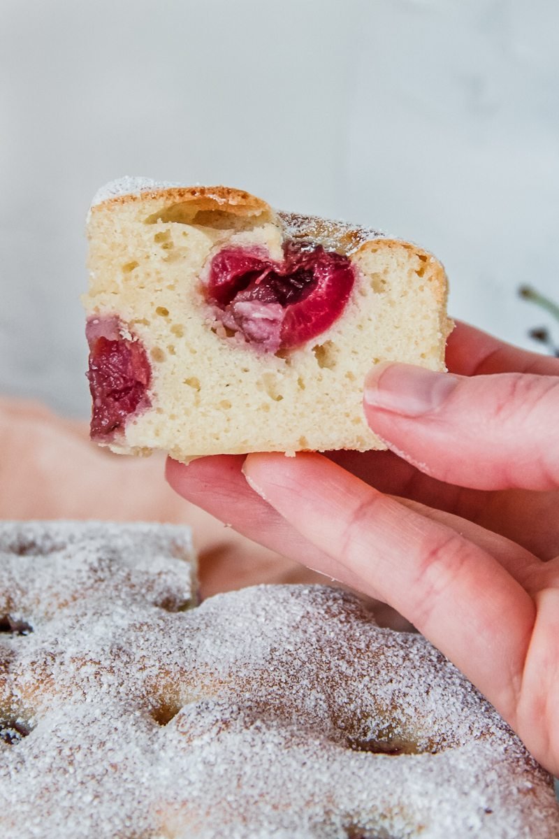 A piece of Cherry Bublanina is held up with the pitted cherries visible in the cake batter.