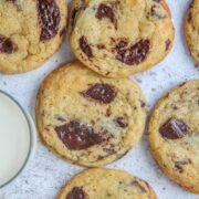 Chocolate chunk cookies lay on a light gray surface with a glass of milk beside.