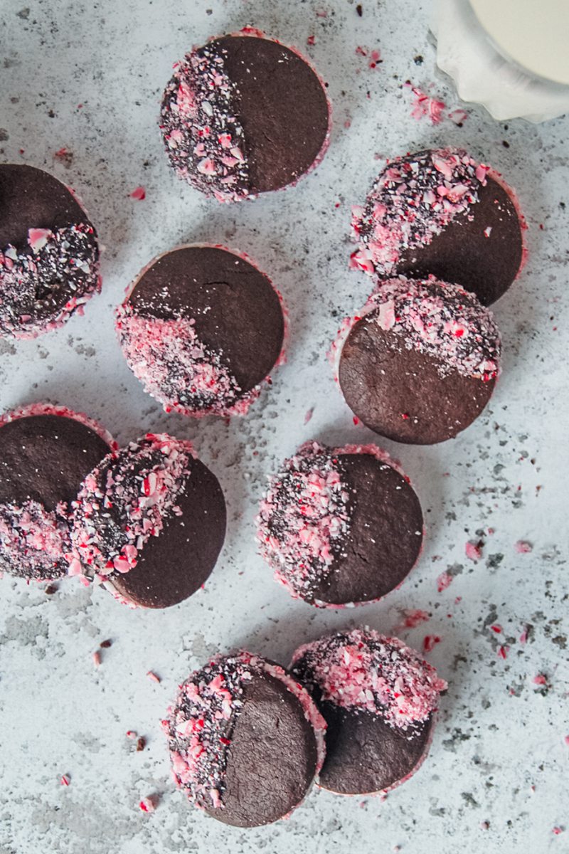 Chocolate peppermint sandwich cookies decorated with crushed candy canes rest on a light gray surface.