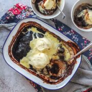 A baked chocolate pudding sits in a metal tin on a gray surface with scoops of melting vanilla ice cream on top with two individual servings beside.