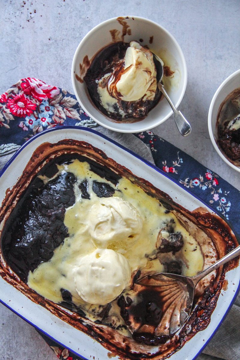 A chocolate self-saucing pudding sits in a metal tin on a gray surface with scoops of melting vanilla ice cream on top with two individual servings beside.