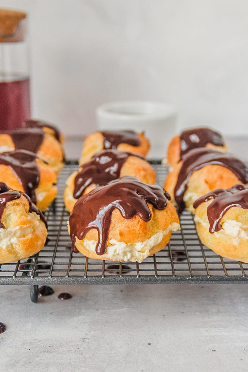 Cream puffs sit on a top of a cooling rack halved and filled with whipped cream and topped with a chocolate sauce on a gray surface.