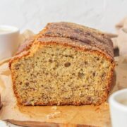 A close up of a slice of classic banana bread sits on a wooden board on a light gray surface.