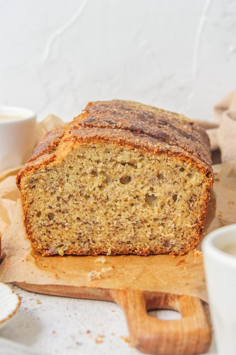 A close up of a slice of classic banana bread sits on a wooden board on a light gray surface.
