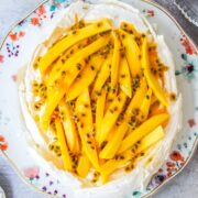 Oval coconut tropical pavlova with passionfruit and mango on top on a floral plate on a gray surface.