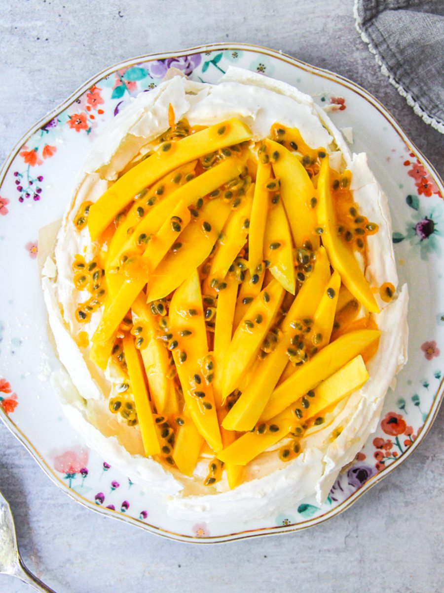 Oval coconut tropical pavlova with passionfruit and mango on top on a floral plate on a gray surface.