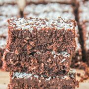 A stack of coconut rough slice bars sit stacked on each other with remaining bars behind on brown parchment paper.