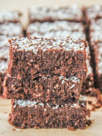 A stack of coconut rough slice bars sit stacked on each other with remaining bars behind on brown parchment paper.