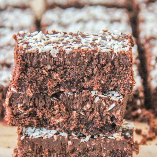 A stack of coconut rough slice bars sit stacked on each other with remaining bars behind on brown parchment paper.