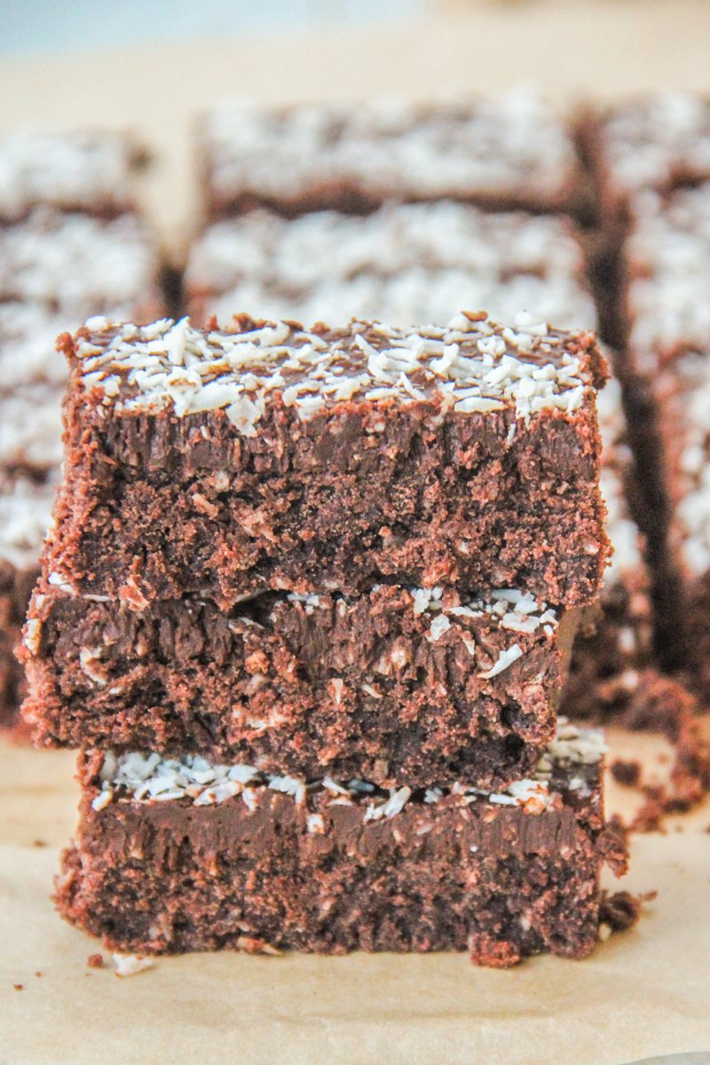 A stack of coconut rough slice bars sit stacked on each other with remaining bars behind on brown parchment paper.