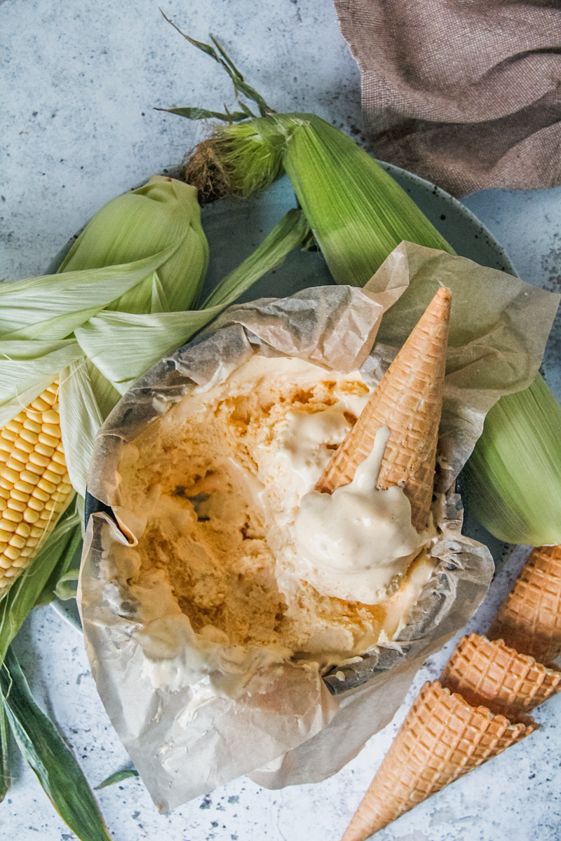 A scoop of corn ice cream in a cone rests on top of a round lined metal tin on a light gray surface with corn cobs beside.