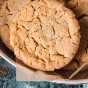 A close up of Cornish Fairing or British Ginger biscuits sitting on a rimmed ceramic plate lined with parchment paper above a green and white surface.