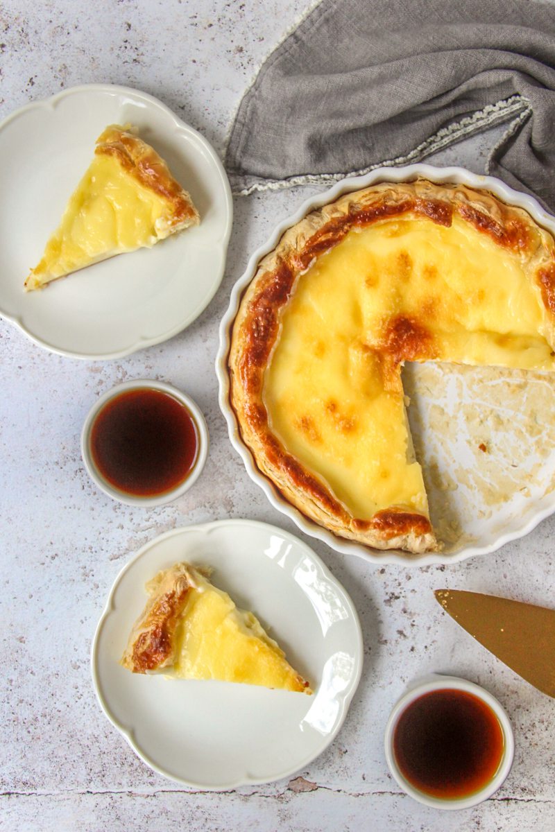 Belgian Cream Pie - Tarte a la creme sits on a light gray surface with a portion remaining in the ceramic baking dish and slices on fluted white ceramic plates.