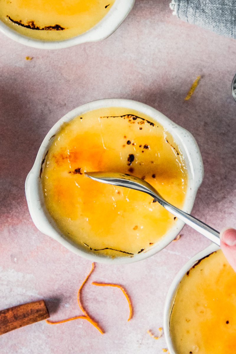 A spoon pierces the caramelized sugar topping of an individual serving of Crema Catalana on a light pink surface.