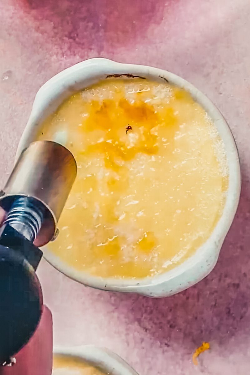 Sugar is caramelized wiith a blowtorch on top of a Crema Catalana in an individual ramekin on a light pink surface.