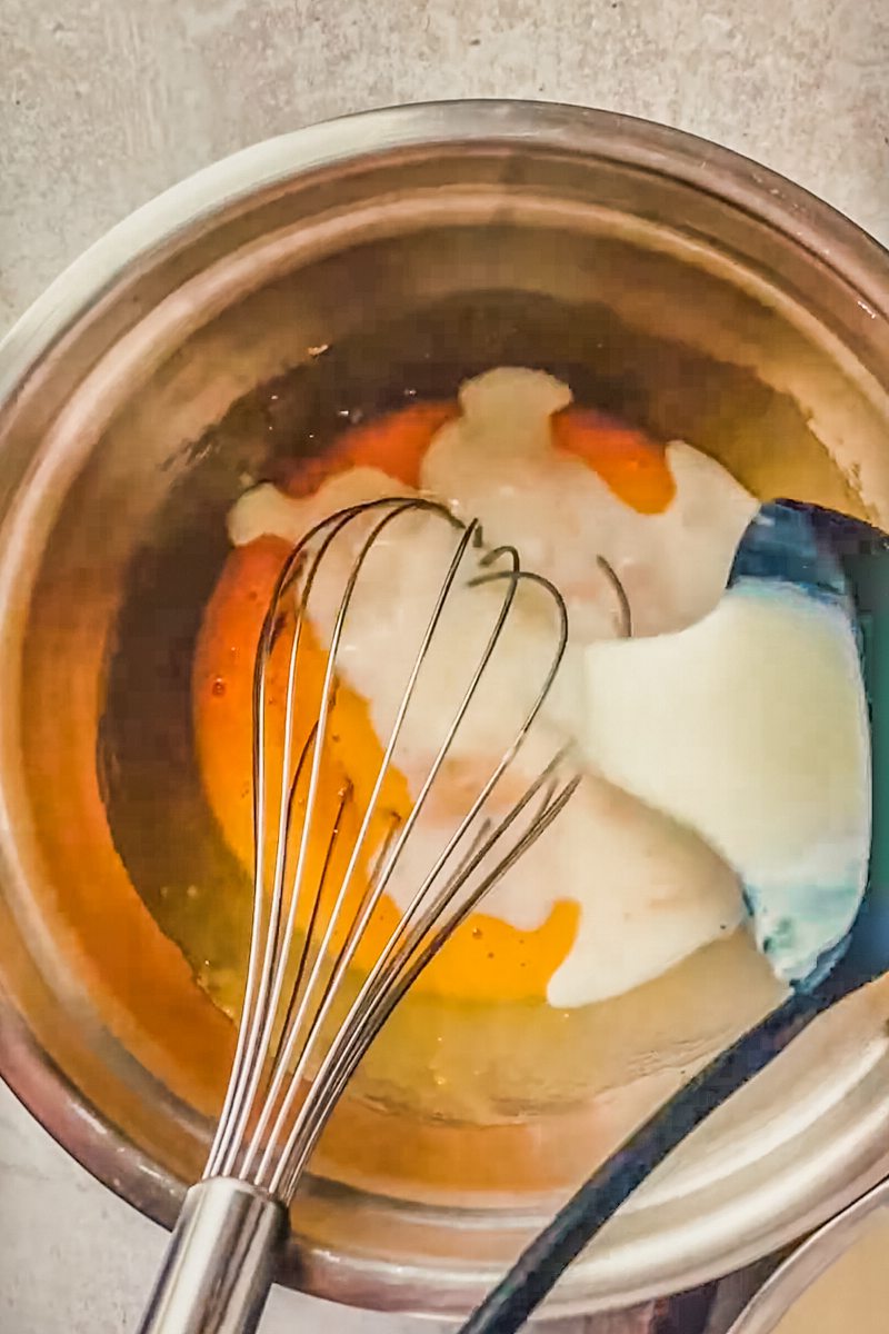 Hot milk is whisked into whisked egg yolks in a stainless steel bowl.