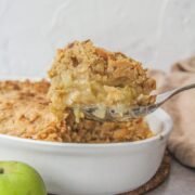 A spoonful of apple fennel crumble sits on a serving spoon above the remaining crumble in a white oval ceramic dish on a gray surface.