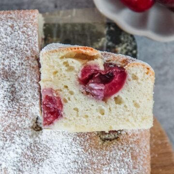 A slice of Czech Bublanina sits on it's side with the light sponge cake texture and halved cherries visible on a wooden board on a light grey background.