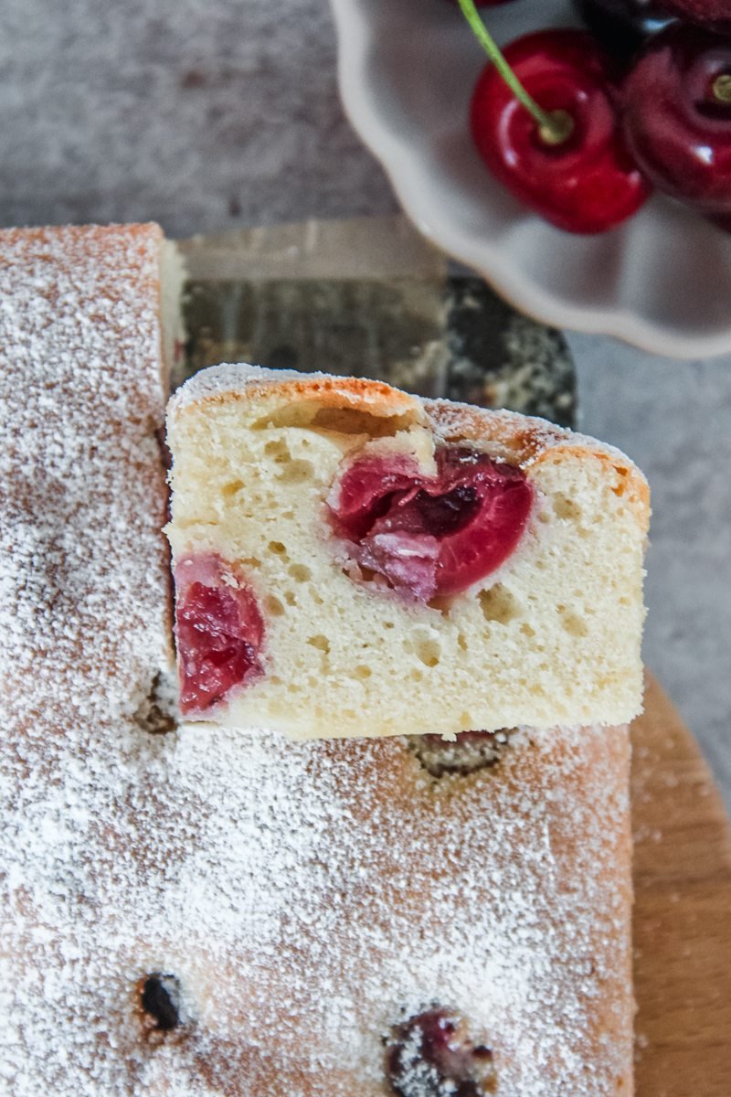 A slice of Czech Bublanina sits on it's side with the light sponge cake texture and halved cherries visible on a wooden board on a light grey background.