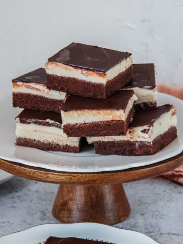 Three layer Czech chocolate cheesecake pieces are stacked on top of each other on a white scalloped plate on a cake stand on a light gray surface.