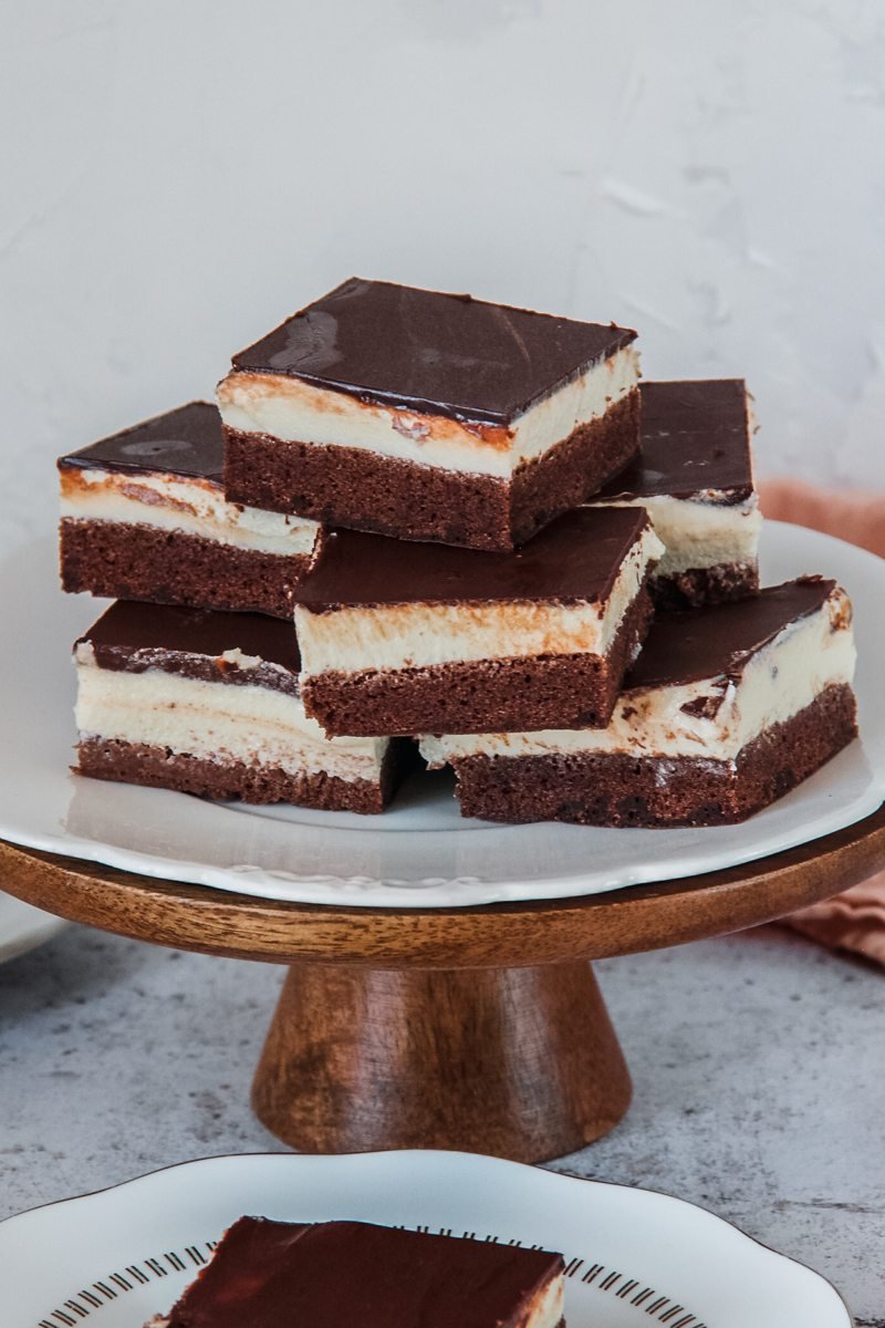 Three layer Czech chocolate cheesecake pieces are stacked on top of each other on a white scalloped plate on a cake stand on a light gray surface.