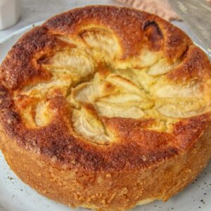 A close up of Dorset Apple Cake sits on a light blue ceramic plate on a grey surface with two small cups of coffee beside.