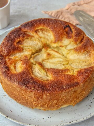 A close up of Dorset Apple Cake sits on a light blue ceramic plate on a grey surface with two small cups of coffee beside.