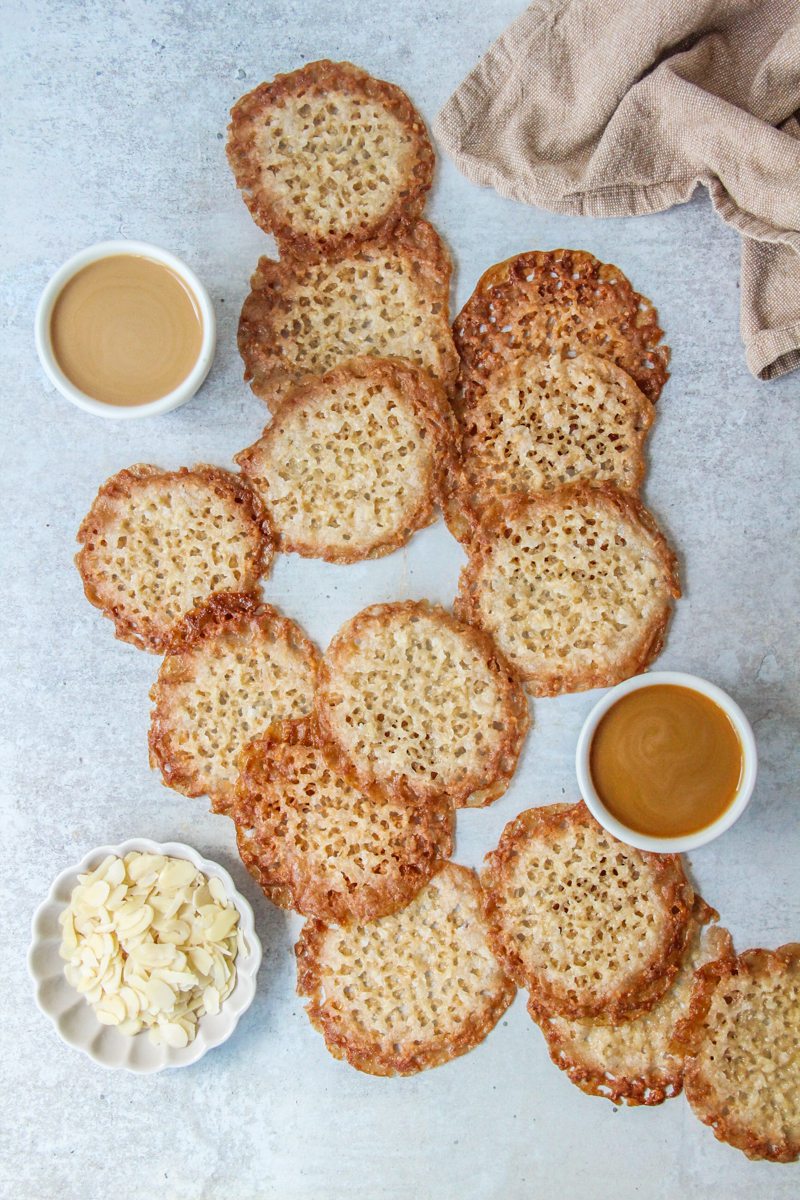 Dutch Almond Lace Biscuits lay on a gray surface with cups of coffee and a small plate of sliced almonds beside.