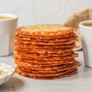 A stack of lace cookies sits on a gray surface with two cups of coffee beside.