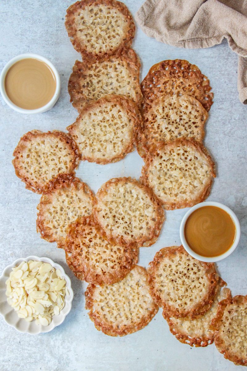 Dutch Kletskoppen lay on a gray surface with cups of coffee and a small plate of sliced almonds beside.