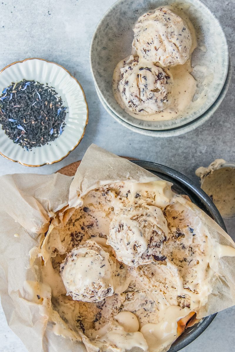 Scoops of earl grey chocolate ice cream sit in both the lined metal tin and a blue grey ceramic bowl with a small plate of earl grey tea beside on a gray surface.