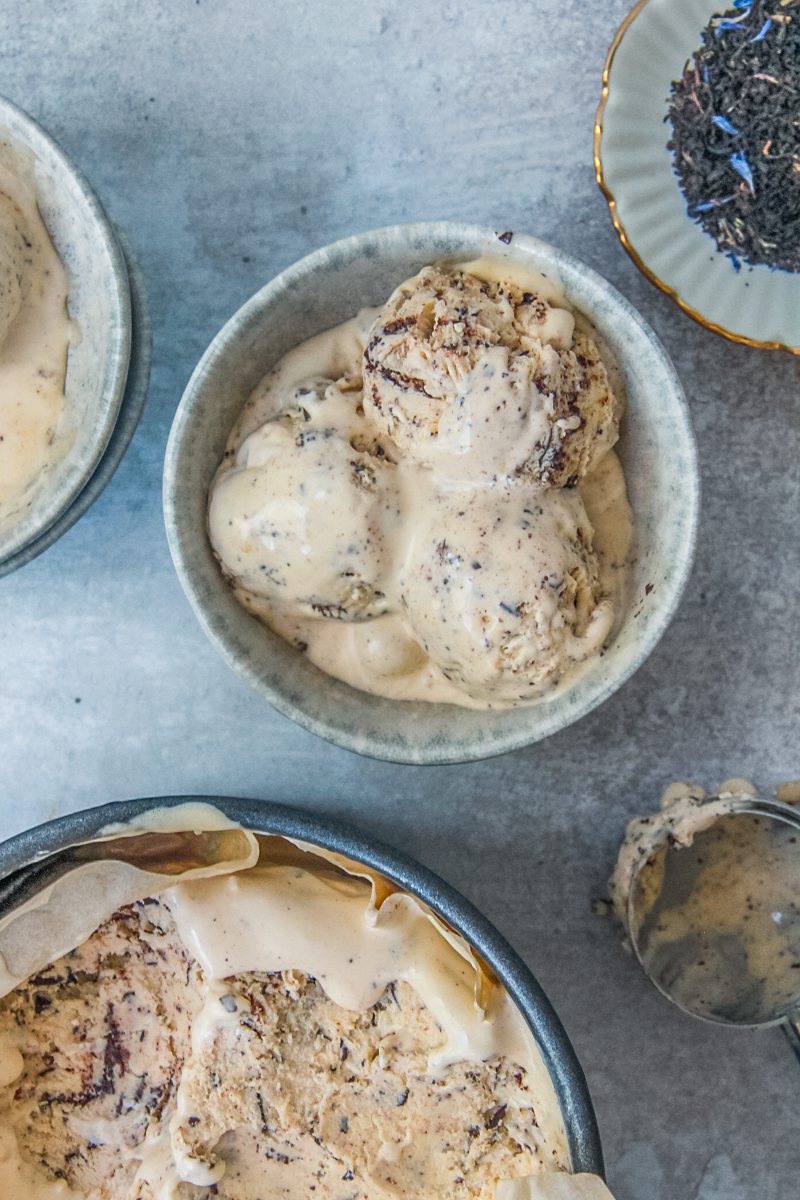 Scoops of earl grey Stracciatella ice cream sit in blue grey ceramic bowls with a small plate of earl grey tea and the tin of ice cream beside on a gray surface.