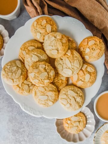 Small golden almond macaroon cookies sit on a fluted white ceramic plate on a gray surface.