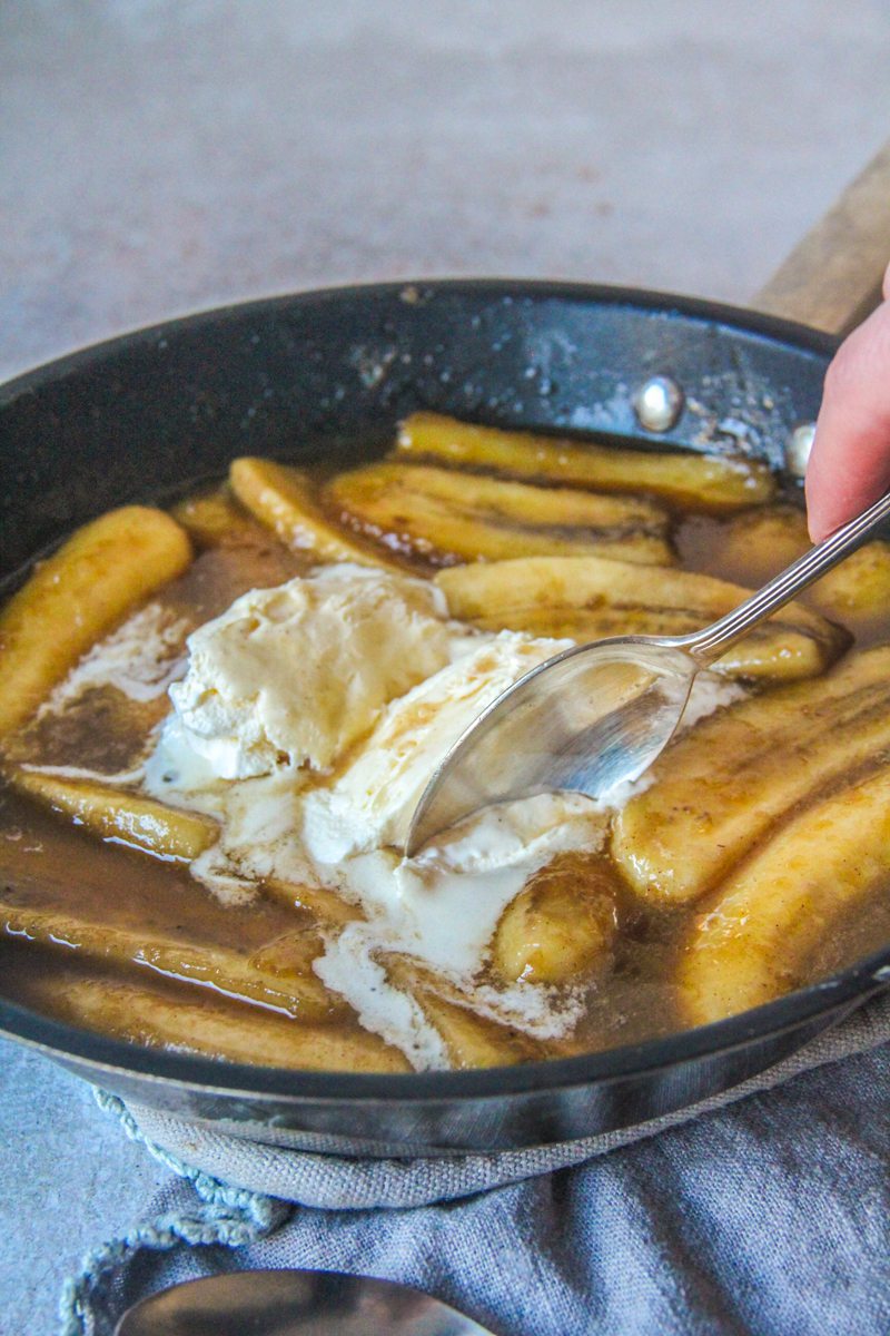 A spoon is dug into a scoop of vanilla ice cream in the middle of banana's foster in a saucepan on a gray surface.