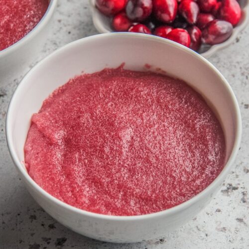 Cranberry pudding sits in white ceramic bowls on a light gray surface with fresh cranberries sitting beside on a small plate.