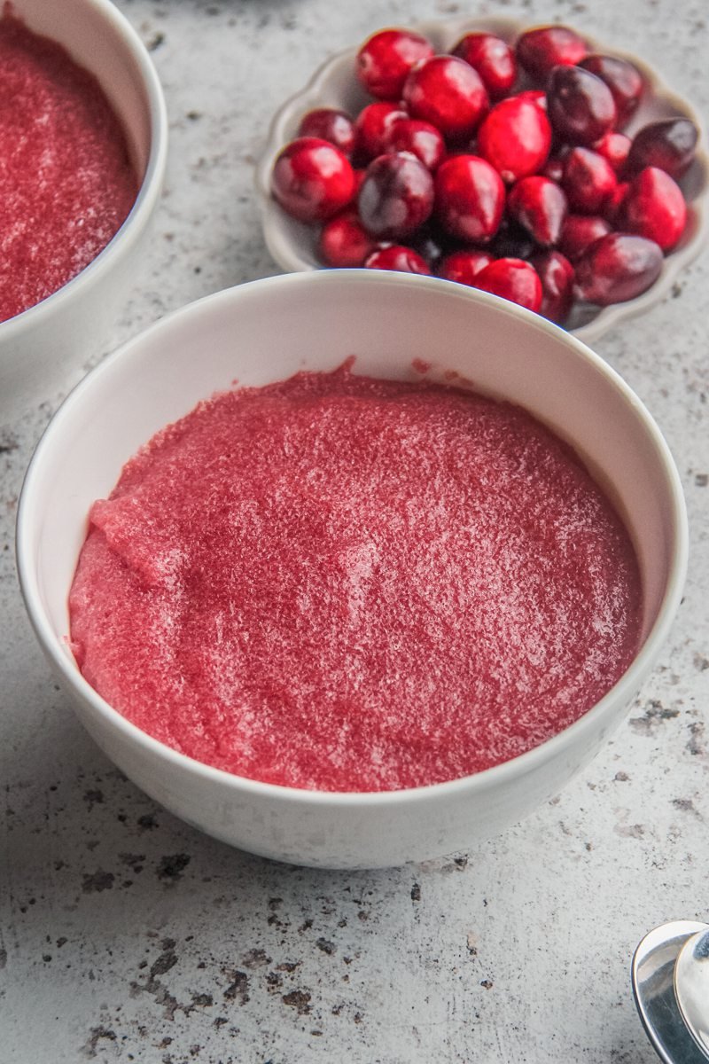 Cranberry pudding sits in white ceramic bowls on a light gray surface with fresh cranberries sitting beside on a small plate.