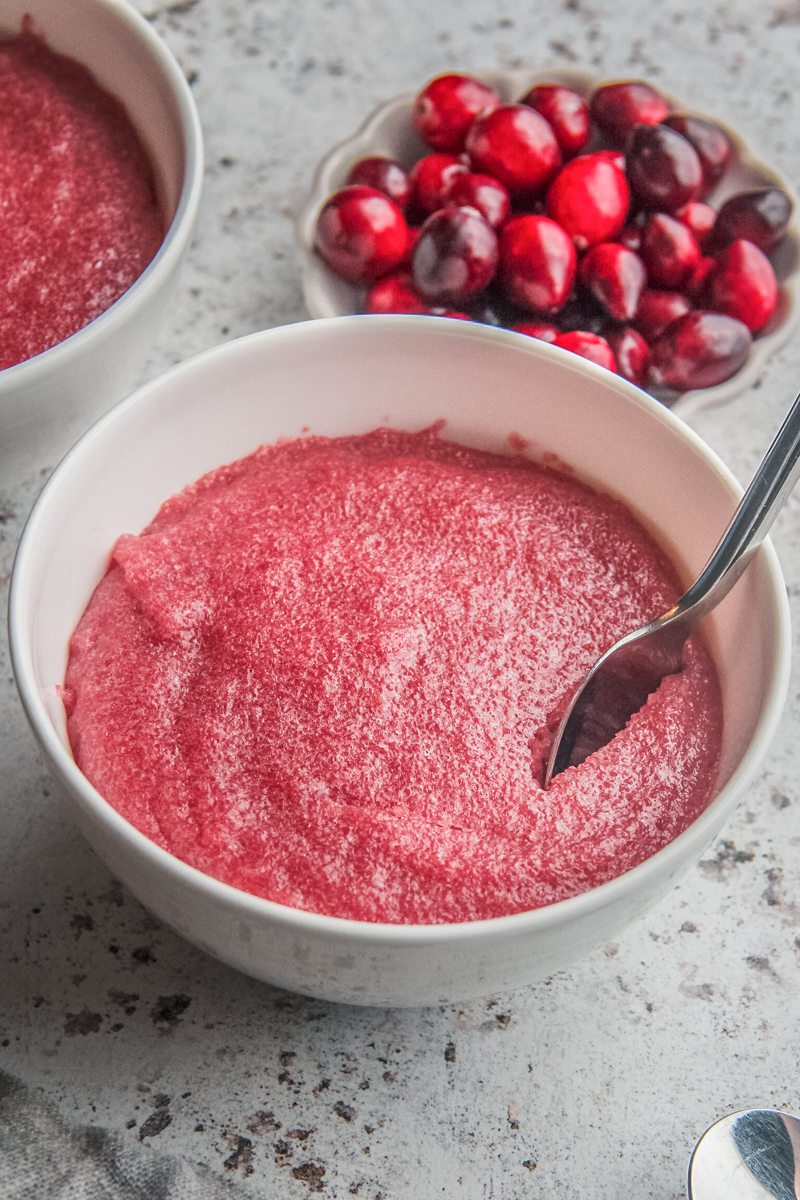 A spoon sinks into Estonian cranberry pudding siting in white ceramic bowls on a light gray surface with fresh cranberries sitting beside on a small plate.