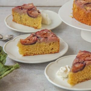 Slices of upside down Fig cake sit on scalloped white ceramic plates on a gray surface with the remaining cake on a stand beside and some dried fig leaves nearby.