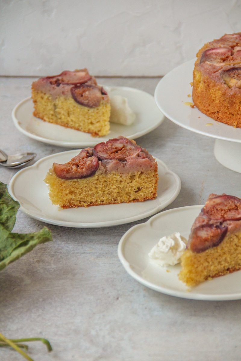 Slices of upside down Fig cake sit on scalloped white ceramic plates on a gray surface with the remaining cake on a stand beside and some dried fig leaves nearby.