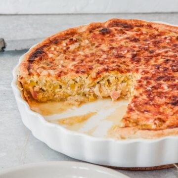 A Flamiche tart with it's leek and bacon interior visible sits in a white ceramic tart dish on a gray surface.