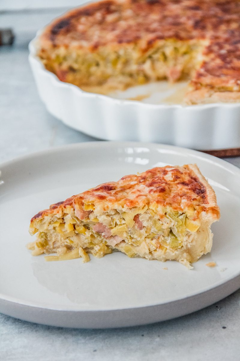 A slice of Flamiche aux Poireaux sits on an individual plate on a gray surface with the remaining tart behind.