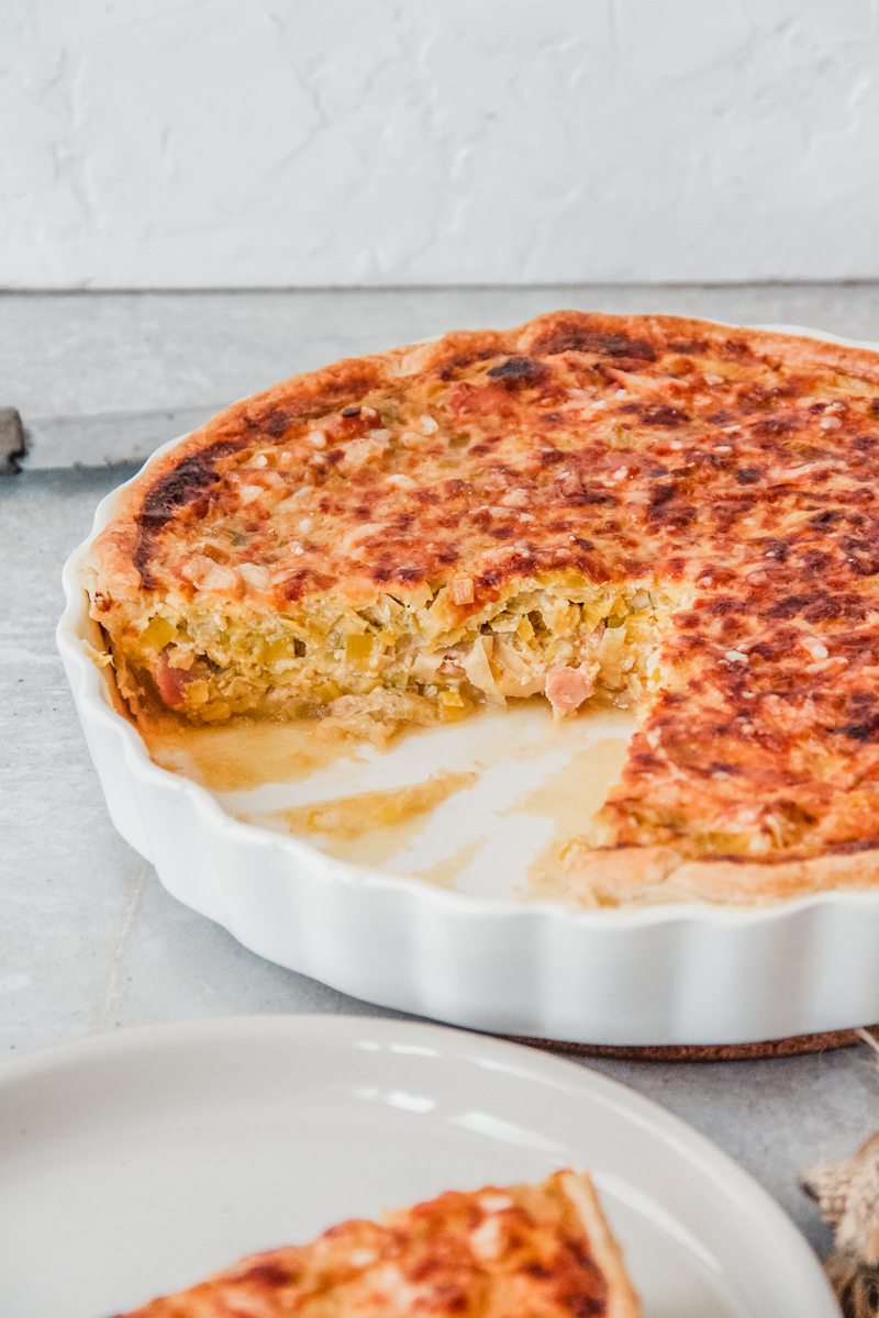 A Flamiche tart with it's leek and bacon interior visible sits in a white ceramic tart dish on a gray surface.